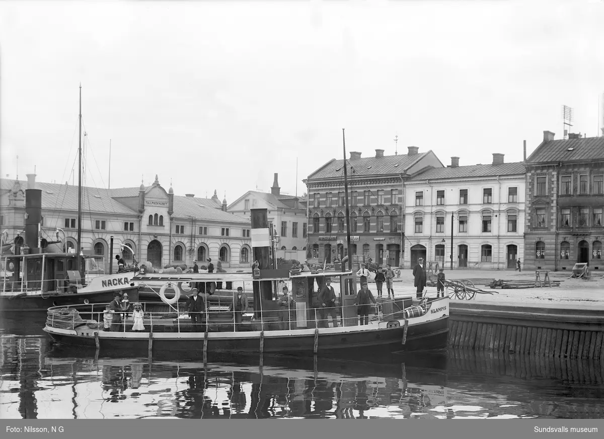 Bogserbåtarna "Klampis" från Klampenborgs sågverk och "Nacka" från Nacka sågverk, ligger förtöjda I "Finska viken" i Sundsvalls hamn. Från vänster Tullpackhuset (tullens kontor). Det ljusa huset till höger byggdes av Handelbanken och där instiftades Sundsvalls första frimurarloge 1886.