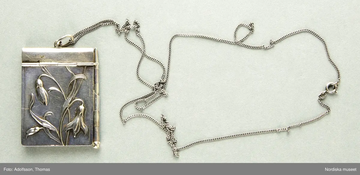 a-c) Ett dansprogram-halsband i jugendstil. a) Dansprogrammetspärm tillverkat i silver, framsidan med reliefdekor av böljande blomsterstjälkar. På dansprogrammets ena långsida är en ögla för fästandet av b) den medföljande lilla smala silverpennan. På dansprogrammets ena kortsida finns en öga där c) en lång silverhalskedja av tunnare pansarmodell är innträd, för praktiskt hängande dansprogrammet runt halsen. Dansprogrammet innehåller flera papperssidor, samtliga med guldkant. På vissa av sidorna står kvinnonamn, vissa endast förnamn andra med fullständigt namn. Sista datumanteckningen är 20/12 1975 och sista anteckningen är Hariette Astrid,

/Cecilia Wallquist 2019-02-21