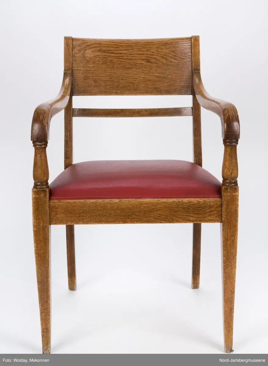 Stol i heltre med stoppet sete med rødt trekk. Stolen har forseggjorte armlener som avsluttes i en myk bue, med dreide detaljer. Ryggstø har en bred plate øverst og en tynnere list under den.