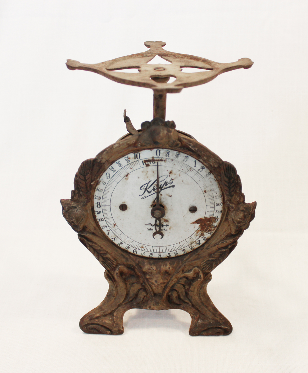 Fjærvekten går til 10 kg og er av merket Krups. Vekten er støpt i jern og blikkplater har blitt brukt på baksiden. 

Fra samlingen etter Ole Gjestvang. 