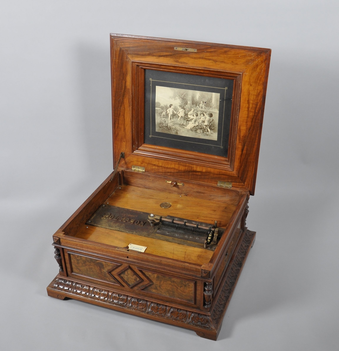 Spilledåse av tre. Spilledåsen har rektangulær form. Står på sokkel med fire bein. Øverste del av sokkel har en borde med palmetter, denne delen har en profilert svai. Selve kassen har dreide pillarer i hjørnene med dekorative kanneleringer. På sidene av kassen er det plater med valbjørk. På framsiden av kassen er det en dekorativ, konsentrisk rombe. Lokket er profilert, med oppstånde midtdel. Midtdelen har svungne sider, med flatskurd av tynne ranker. På toppen av midtdelen, er det intarsia med motiv av lyse blomster på mørk bakgrunn. Treverket er lakkert. 
Lokket er hengslet, det er et litografi/trykk på innside av lokk med motiv av barn som spiller instrumenter.