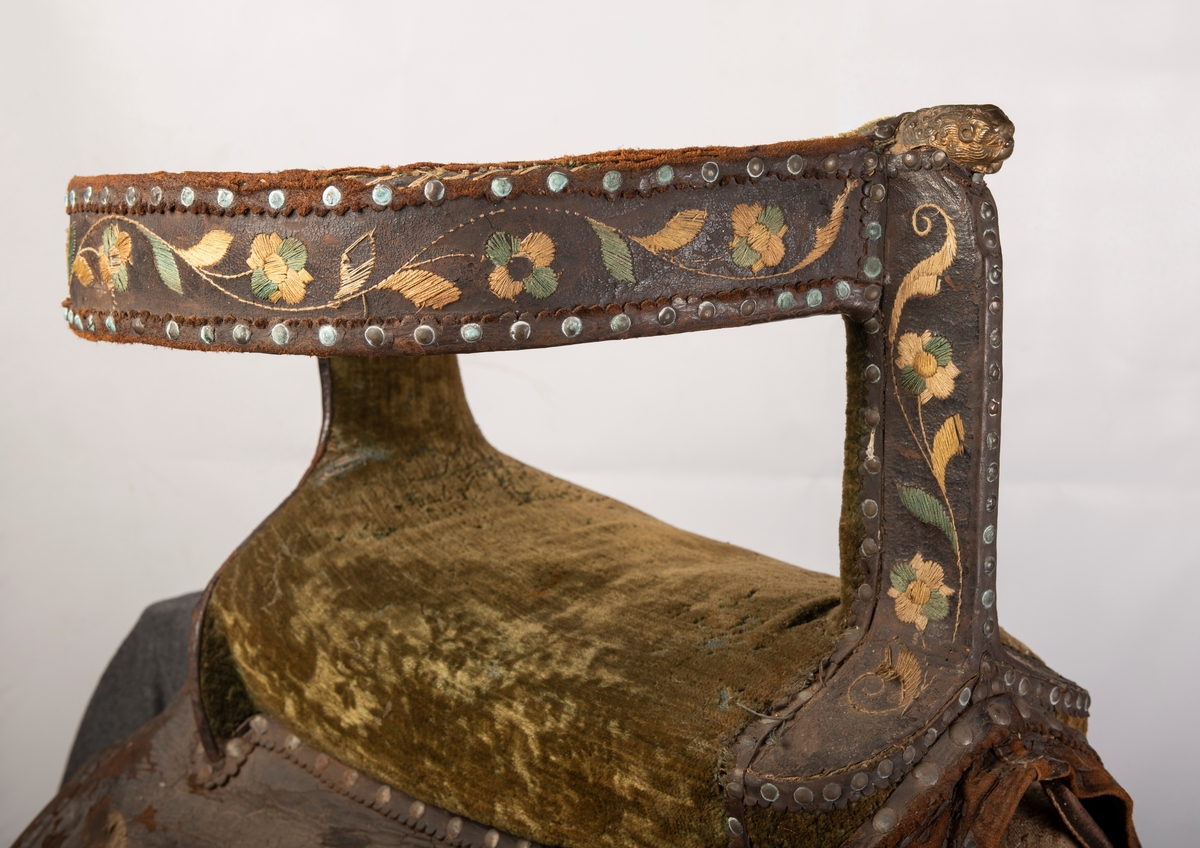 Brodert lær. Grønn mønstret  fløyel i setet og på armlene. Messinghode på fremre del av rygglenet. Initialene KDS og årstallet1773 brodert på. Løs narv på kappene, redrot.