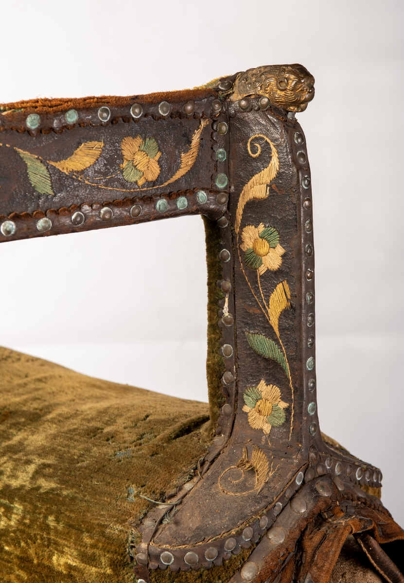 Brodert lær. Grønn mønstret  fløyel i setet og på armlene. Messinghode på fremre del av rygglenet. Initialene KDS og årstallet1773 brodert på. Løs narv på kappene, redrot.