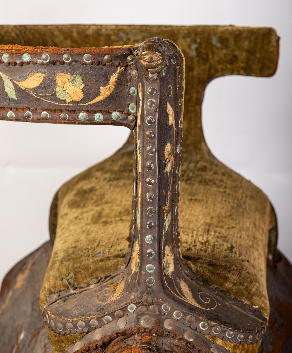 Brodert lær. Grønn mønstret  fløyel i setet og på armlene. Messinghode på fremre del av rygglenet. Initialene KDS og årstallet1773 brodert på. Løs narv på kappene, redrot.