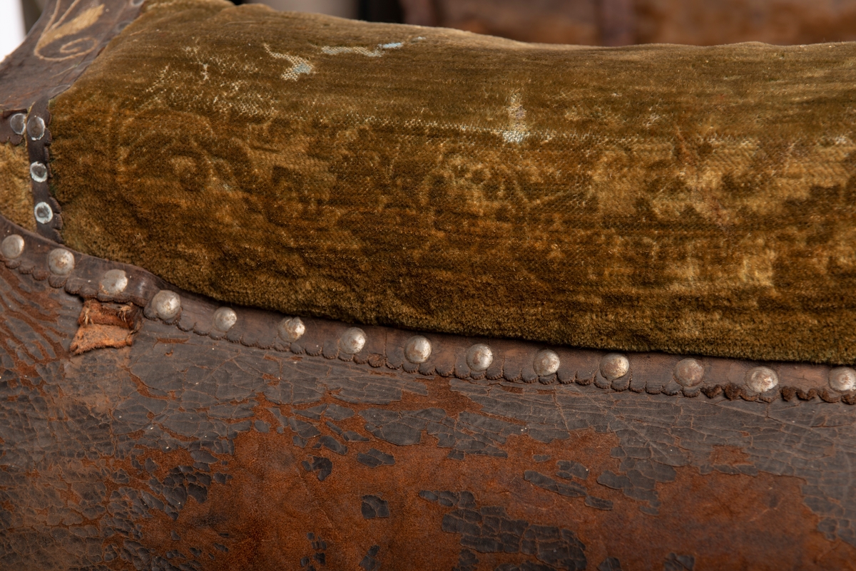 Brodert lær. Grønn mønstret  fløyel i setet og på armlene. Messinghode på fremre del av rygglenet. Initialene KDS og årstallet1773 brodert på. Løs narv på kappene, redrot.