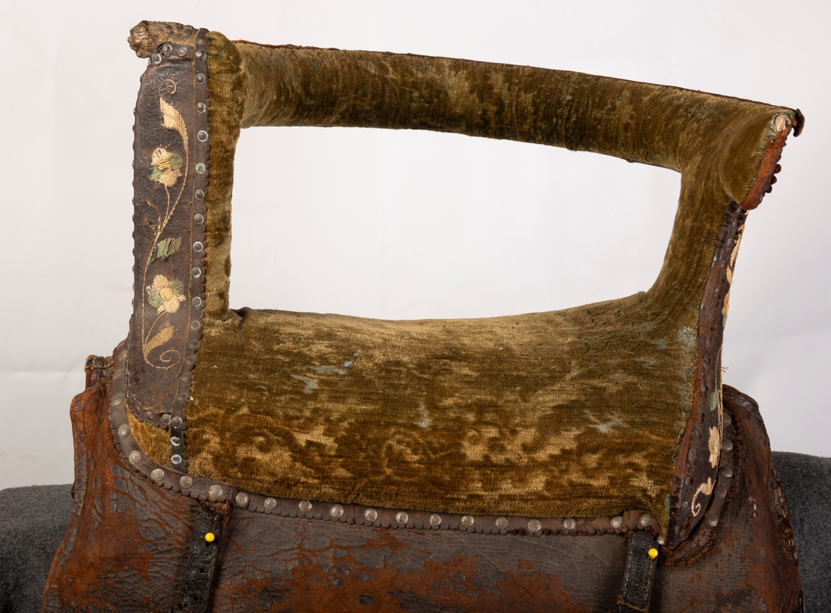 Brodert lær. Grønn mønstret  fløyel i setet og på armlene. Messinghode på fremre del av rygglenet. Initialene KDS og årstallet1773 brodert på. Løs narv på kappene, redrot.