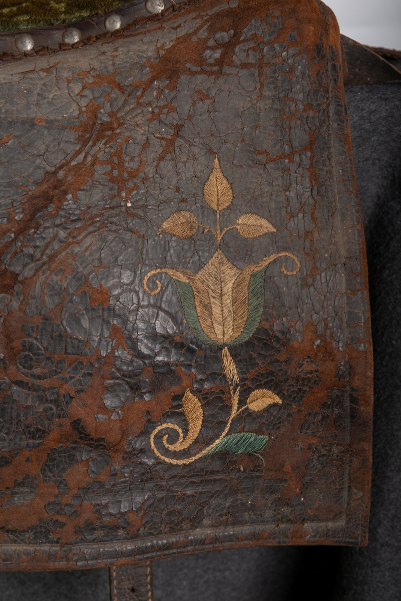 Brodert lær. Grønn mønstret  fløyel i setet og på armlene. Messinghode på fremre del av rygglenet. Initialene KDS og årstallet1773 brodert på. Løs narv på kappene, redrot.