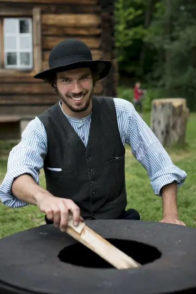 En museumsvert kledd som skogfinne smiler mens han fyrer bål ved Ampiansbråten på Norsk Folkemuseum.