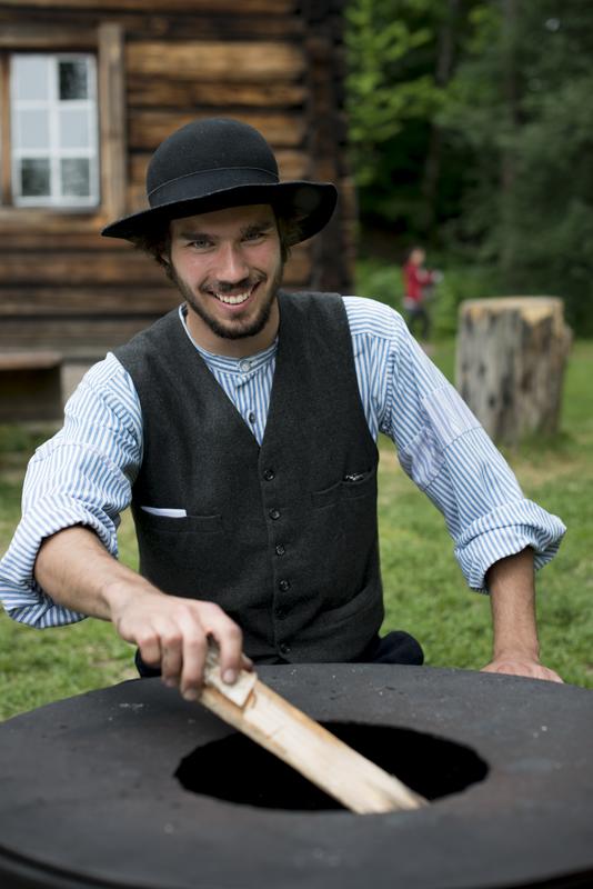 En museumsvert kledd som skogfinne smiler mens han fyrer bål ved Ampiansbråten på Norsk Folkemuseum.