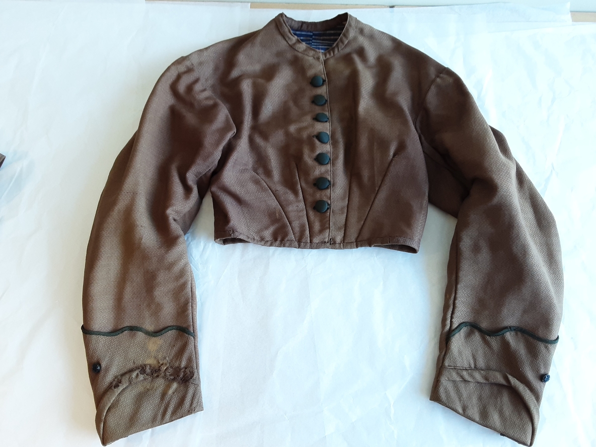 Todelt kjole i tynn brokade med blomstermønster innvevd på skjørtet. Kjolelivet er helfòret med to ulike stoffer. ermene er sydd i for hånd og sårkantene kastet over for hånd. stikningssømmer er sydd på maskin. Sidesømmer og innsnitt med lett spiling likeså. Kjolelivet lukkes foran med stofftrukne knapper og to hekter nederst. Nederst på ermene er det pyntet med oppbrett med grønne bånd, kapper og en effekt i kjolens stoff. Ermene er skåret i to deler.
Skjørtet er foldet foran med splitt i fold på venstre side som lukkes med hekter på to steder. Nederst på innsiden er det skoningskant og slitesnor. Bak i livet er det et felt på 6 cm med striping.
Belte med hektelukning under sløyfe som har sittet bak.
Kjøpt stoff. Muligens brukt som modell for stakk til Sør-Østerdalsbunaden.