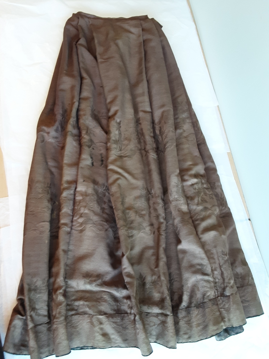 Todelt kjole i tynn brokade med blomstermønster innvevd på skjørtet. Kjolelivet er helfòret med to ulike stoffer. ermene er sydd i for hånd og sårkantene kastet over for hånd. stikningssømmer er sydd på maskin. Sidesømmer og innsnitt med lett spiling likeså. Kjolelivet lukkes foran med stofftrukne knapper og to hekter nederst. Nederst på ermene er det pyntet med oppbrett med grønne bånd, kapper og en effekt i kjolens stoff. Ermene er skåret i to deler.
Skjørtet er foldet foran med splitt i fold på venstre side som lukkes med hekter på to steder. Nederst på innsiden er det skoningskant og slitesnor. Bak i livet er det et felt på 6 cm med striping.
Belte med hektelukning under sløyfe som har sittet bak.
Kjøpt stoff. Muligens brukt som modell for stakk til Sør-Østerdalsbunaden.