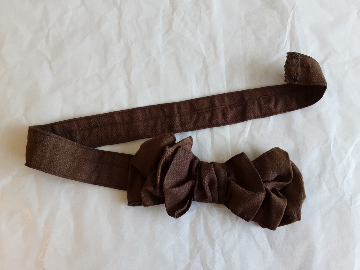 Todelt kjole i tynn brokade med blomstermønster innvevd på skjørtet. Kjolelivet er helfòret med to ulike stoffer. ermene er sydd i for hånd og sårkantene kastet over for hånd. stikningssømmer er sydd på maskin. Sidesømmer og innsnitt med lett spiling likeså. Kjolelivet lukkes foran med stofftrukne knapper og to hekter nederst. Nederst på ermene er det pyntet med oppbrett med grønne bånd, kapper og en effekt i kjolens stoff. Ermene er skåret i to deler.
Skjørtet er foldet foran med splitt i fold på venstre side som lukkes med hekter på to steder. Nederst på innsiden er det skoningskant og slitesnor. Bak i livet er det et felt på 6 cm med striping.
Belte med hektelukning under sløyfe som har sittet bak.
Kjøpt stoff. Muligens brukt som modell for stakk til Sør-Østerdalsbunaden.