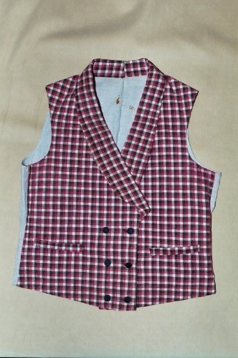 Vest i rutet bomullslerret i fargene rødt, olivengrønt/grønnnbrunt og ubleiket hvitt. Fôr og rygg i linlerret. Vesten er dobbeltspent med svarte, stofftrukne knapper. Den har sjalskrage og spensel i ryggen. Plagget er helt håndsydd. På forstykkene er det to lett skråstilte lommer og vesten buer ned midt foran.