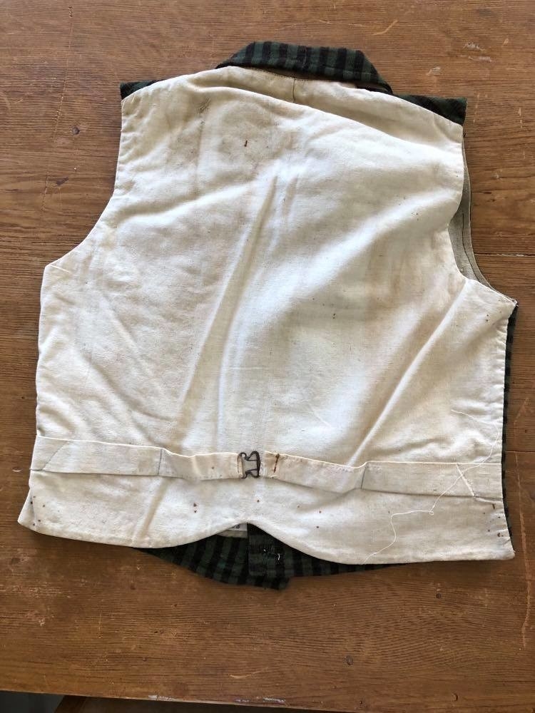 Dobbeltspent vest i ullverken, stripet i grønt og svart (sauesvart) med brun renning. Dobbel rygg i bomullslerret. Halvlinslerret (bomull/stry) i fôret i framstykkene. Vesten har fire knapphull sydd med kamgarnsullgarn og svarte knapper trukket med svart bomullssateng med brun renning. Plagget har nedbrettet krage og slag/reverser som kan kneppes mot bolen. Knapp og knapphull. Vesten buer ned midt framme og har svakt skråstilte lommer. Vattering av stry i framstykkene. I ryggen buer vesten opp midt bak. Den er vrangsydd langs ytterkantene og stukket ned med maskin, 0,8 cm fra kanten. (Registrering ved Bunad- og folkedraktrådet/Norsk institutt for bunad og folkedrakt v/Jon Fredrik Skauge, 2005, revidert v/Bjørn Sverre Hol Haugen, 2020.)