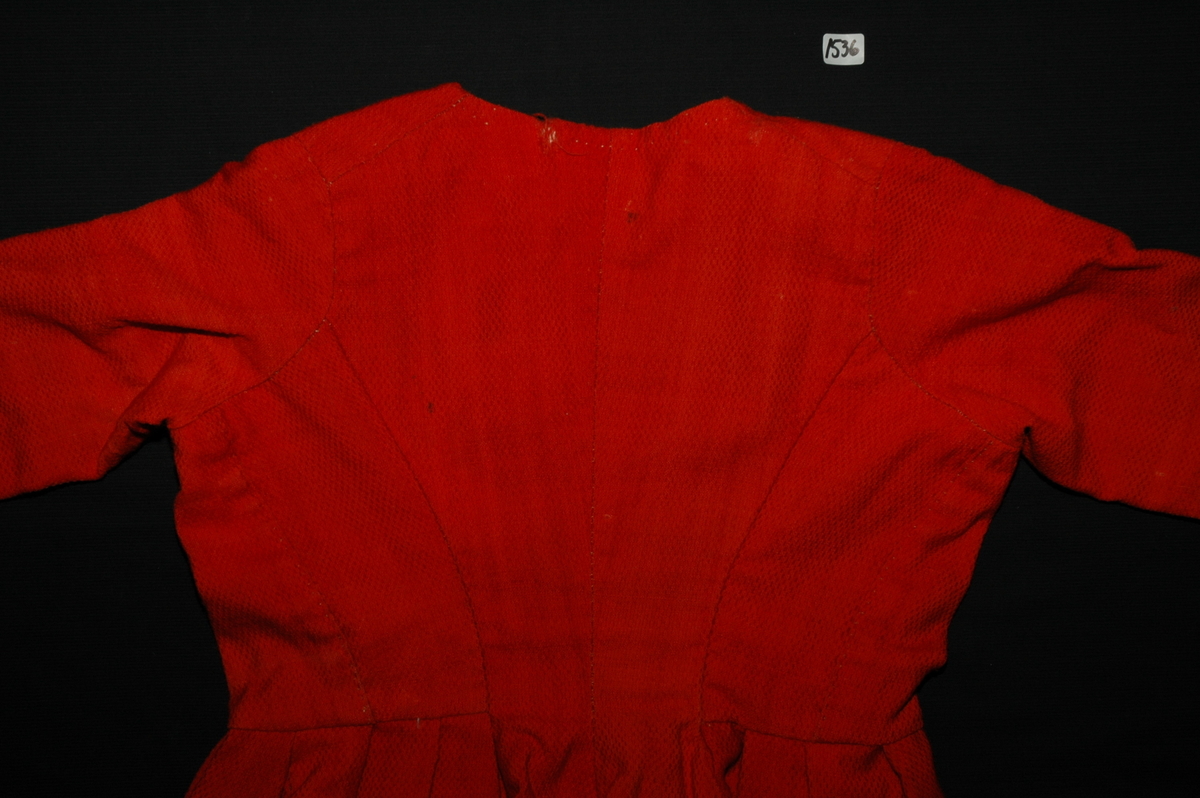 Trøye av rødt ullstoff med linfôr. Stoffet er av kamgarn og ble antageligvis solgt under betegnelsen everlasting på 1700-tallet. Produksjonssted er antageligvis England. Stoffet er i toskaft binding med flotteringer over tre tråder som gir mønstereffekt. Trøya har vært lukket med ni hekter og maljer midt framme. Snitt: Framstykkene er avdelt i midjen med kappe. Ryggstykkene går helt ned i skjøten. Kappa er foldelagt med sju folder (fra 10 cm midt framme). Foldene er 3-3,5 cm brede og 1,5 cm dype. Bua ryggsømmer sydd med spilesøm. Ryggsømmen midt bak er sydd med attersting hver for seg, men festet sammen i fôr og hovedstoff på noen steder. Det er to innsnitt under hvert herme. Ytterkantene er faldet med faldeprikkesting. Det er ekstra prikkestingsrad ved halsringing (1 cm fra ytterkant) og ved hektestillinga omlag 2 cm fra ytterkanten framme. Kappa er sydd til bolen med attersting (tre lag stoff). Fôret i bolen er bretta og lagt til og sydd ned i fôret til kappa. Kappa skrår bakover midt framme. Trange ermer sydd sammen med attersting. Sømmonnet er lagt mot bolen og fôret i bolen er lagt mot ermet og sydd til med faldesting. Skinnhemper til opphenging. Ensømserme med albuestnitt. På unerermene er det et innsnitt. Underermssømmer er sydd med attersting, stoff og fôr hver for seg (festet sammen på enkelte steder). Innsnittene er sydd med stoff og fôr sammen og lagt til en side og stukket ned, faldet til side. (Fra regsitrering ved Bunad- og folkedraktrådet/Norsk institutt for bunad og folkedrakt v/Jon Fredrik Skauge i 2005, revidert v/Bjørn Sverre Hol Haugen 2020). 