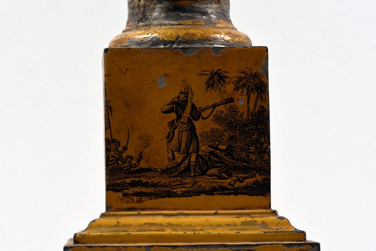 En av ett par fotogenlampor med gulmålad fot i plåt. På målningen finns tryckta stridsscener samt dekor med gotiska valv i svart. Foten är i form av en pelare på kvadratisk bas. Fotogenbehållaren är av glas och brännaren av mässing. Lampkupan är av opalfärgat glas med inslipad blomsterdekor. Högst upp ett skydd sytt i cremefärgat sidentyg, i form av en blomma.