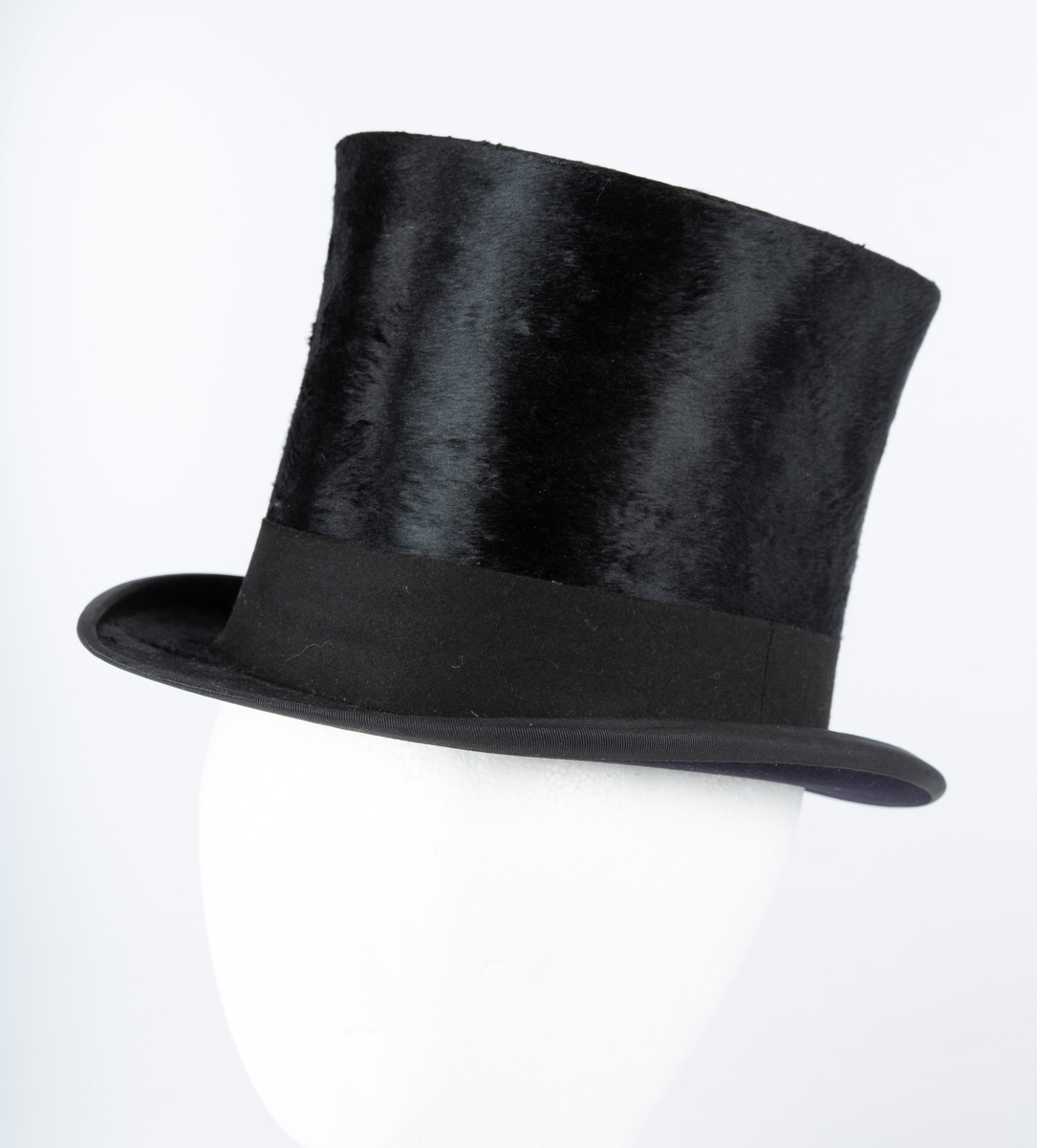 Oval hatt, rett pull med flat topp, flosset stoff, matt hattebånd. Flar brem forran og bak, oppbøyet sidekant. Foret med brun ripssilke. Svettekant i skinn.
Inni hatten et visittkort  med påskrift J. Schaan Theiste, Burauchef

 største LlxB: 30x24  Innvendig mål: L 20  B 15  H 15