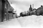 Vintermotiv med stora mängder snö på Brahegatan. Trottoaren till vänster är skottad så snövallarna ligger till höger - mitt i gatan?
Närmast i bild fastigheter med butiksskyltar 