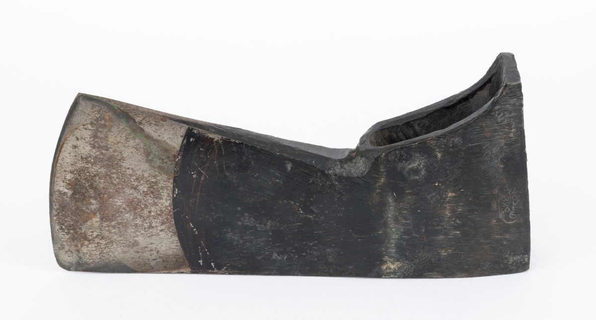 Uskjeftet øks fra produsenten O. Mustad & Søn på Gjøvik i Oppland. Øksehodet er 21,1 centimeter høyt, målt vinkelrett fra midten av den noenlunde plane nakkeflata ned mot den fremre delen av egglinja. Den nederste delen av øksebladet er 9,0 centimeter bred. Her er målet tatt i rett linje mellom den konvekse eggens ytterkanter. Den fremre tverrenden på øksehodet er noenlunde rettlinjet. På «baksida» skrår bladet noe innover mot et punkt like under «øyet» (skafthullet), der bredden er 5,7 centimeter. På denne sida øker bredden på «kjakene» mot den bakre enden av nakkepartiet. Hammerflata på denne delen av øksa er 9,6 centimeter langt og om lag 3,1 centimeter bredt bakerst, 2,8 i den fremre enden. På den høyre sida av øksenakken (sett i øksebrukerens perspektiv) finnes et svakt preget ovalt stempel med teksten «MUSTAD ØKS». Den øvre delen av øksehodet er svartlakkert, mens ei cirka 5,5 centimeter bred sone nederst på bladet er stålblank.