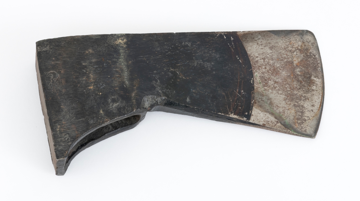 Uskjeftet øks fra produsenten O. Mustad & Søn på Gjøvik i Oppland. Øksehodet er 21,1 centimeter høyt, målt vinkelrett fra midten av den noenlunde plane nakkeflata ned mot den fremre delen av egglinja. Den nederste delen av øksebladet er 9,0 centimeter bred. Her er målet tatt i rett linje mellom den konvekse eggens ytterkanter. Den fremre tverrenden på øksehodet er noenlunde rettlinjet. På «baksida» skrår bladet noe innover mot et punkt like under «øyet» (skafthullet), der bredden er 5,7 centimeter. På denne sida øker bredden på «kjakene» mot den bakre enden av nakkepartiet. Hammerflata på denne delen av øksa er 9,6 centimeter langt og om lag 3,1 centimeter bredt bakerst, 2,8 i den fremre enden. På den høyre sida av øksenakken (sett i øksebrukerens perspektiv) finnes et svakt preget ovalt stempel med teksten «MUSTAD ØKS». Den øvre delen av øksehodet er svartlakkert, mens ei cirka 5,5 centimeter bred sone nederst på bladet er stålblank.