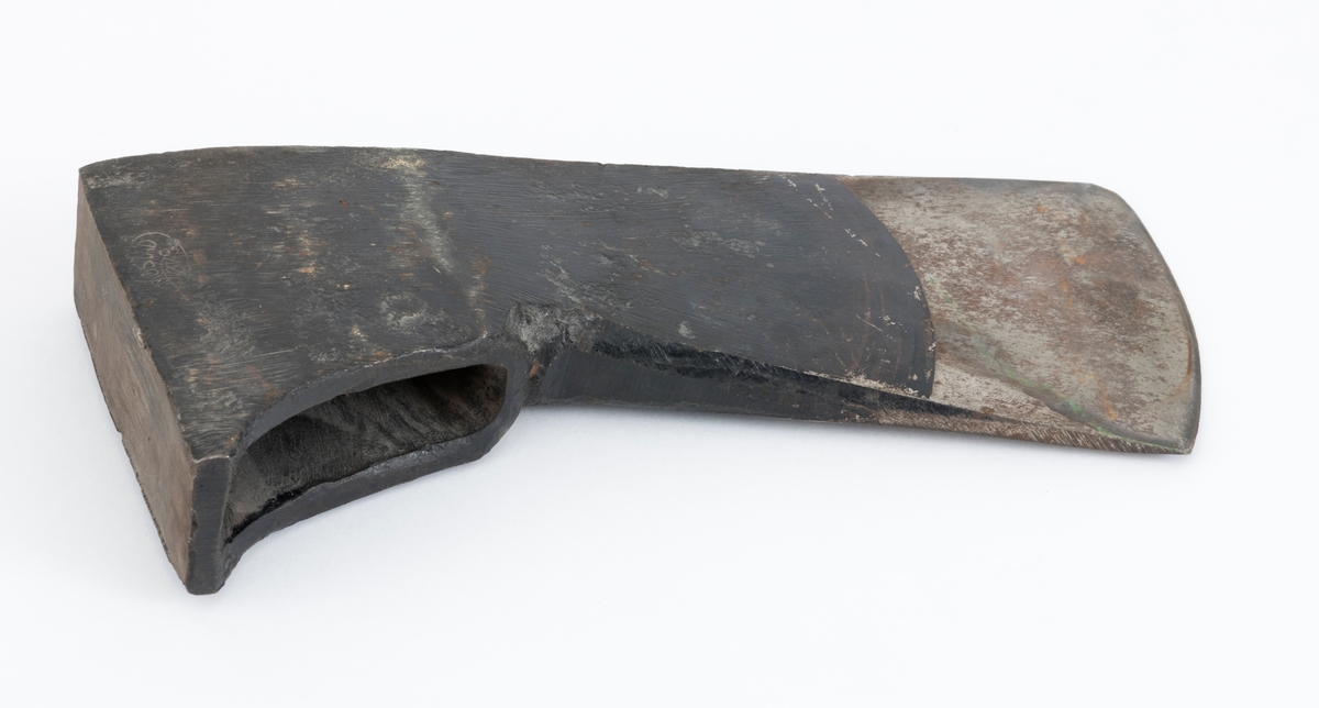 Uskjeftet øks fra produsenten O. Mustad & Søn på Gjøvik i Oppland. Øksehodet er 21,1 centimeter høyt, målt vinkelrett fra midten av den noenlunde plane nakkeflata ned mot den fremre delen av egglinja. Den nederste delen av øksebladet er 9,0 centimeter bred. Her er målet tatt i rett linje mellom den konvekse eggens ytterkanter. Den fremre tverrenden på øksehodet er noenlunde rettlinjet. På «baksida» skrår bladet noe innover mot et punkt like under «øyet» (skafthullet), der bredden er 5,7 centimeter. På denne sida øker bredden på «kjakene» mot den bakre enden av nakkepartiet. Hammerflata på denne delen av øksa er 9,6 centimeter langt og om lag 3,1 centimeter bredt bakerst, 2,8 i den fremre enden. På den høyre sida av øksenakken (sett i øksebrukerens perspektiv) finnes et svakt preget ovalt stempel med teksten «MUSTAD ØKS». Den øvre delen av øksehodet er svartlakkert, mens ei cirka 5,5 centimeter bred sone nederst på bladet er stålblank.