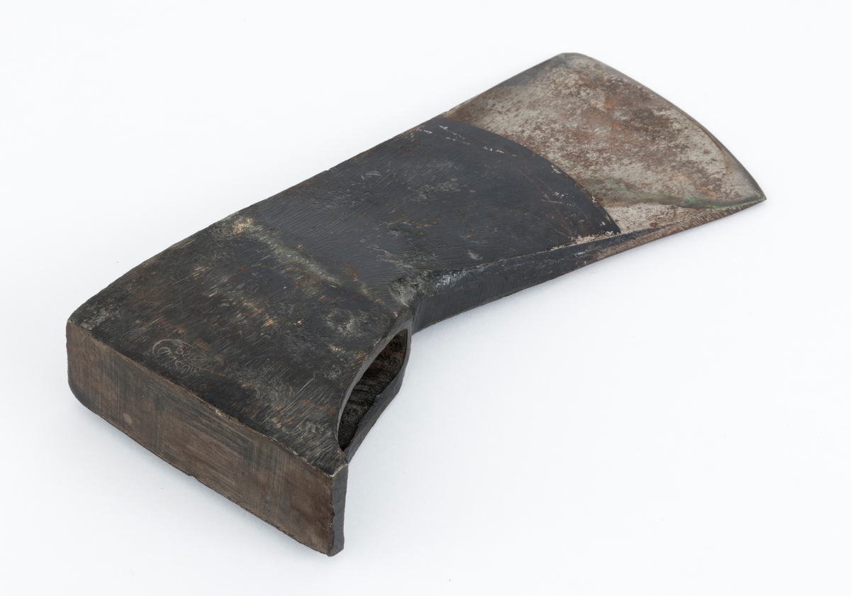 Uskjeftet øks fra produsenten O. Mustad & Søn på Gjøvik i Oppland. Øksehodet er 21,1 centimeter høyt, målt vinkelrett fra midten av den noenlunde plane nakkeflata ned mot den fremre delen av egglinja. Den nederste delen av øksebladet er 9,0 centimeter bred. Her er målet tatt i rett linje mellom den konvekse eggens ytterkanter. Den fremre tverrenden på øksehodet er noenlunde rettlinjet. På «baksida» skrår bladet noe innover mot et punkt like under «øyet» (skafthullet), der bredden er 5,7 centimeter. På denne sida øker bredden på «kjakene» mot den bakre enden av nakkepartiet. Hammerflata på denne delen av øksa er 9,6 centimeter langt og om lag 3,1 centimeter bredt bakerst, 2,8 i den fremre enden. På den høyre sida av øksenakken (sett i øksebrukerens perspektiv) finnes et svakt preget ovalt stempel med teksten «MUSTAD ØKS». Den øvre delen av øksehodet er svartlakkert, mens ei cirka 5,5 centimeter bred sone nederst på bladet er stålblank.