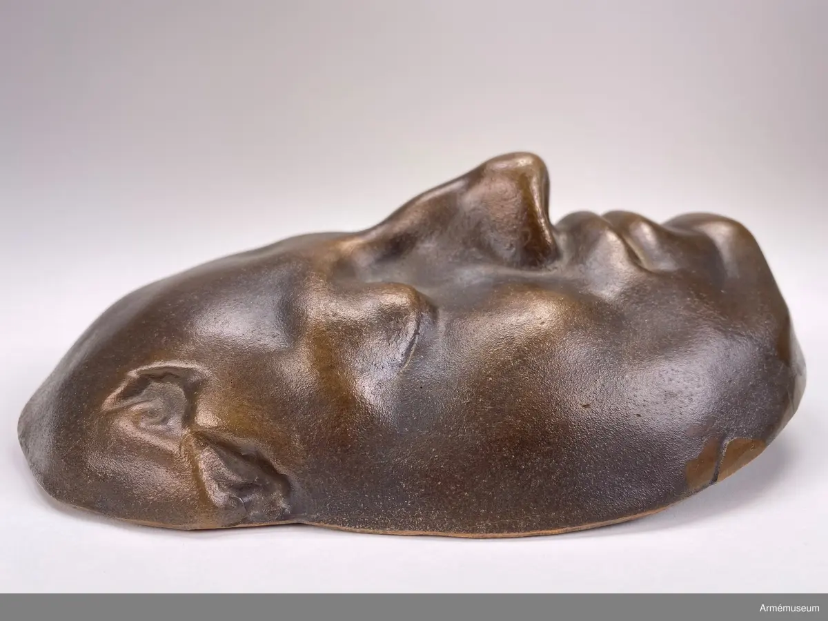 Bronsmask föreställande den döde Karl XII.

Enl styresman Olle Cederlöfs anteckningar med anledning av gåva AM.066063 (AM 73/475) är detta inte en dödsmask. 

"Proportionerna i ansiktet stämmer inte med kraniets proportioner enl 1918 års gravöppning - utan en av den schweiziske medaljgravören Hedlinger gjord frihandsmodellering inför konungens lik på Karlberg 1718. Hedlingers modell nu endast avsedd att ligga till grund för hans uppdrag: Att modellera en reliefbild för konungens mynt och minnespenningar. Hedlinger anlände till Sverige 1718-. Konungen hade då avlidit och Hedlinger hade aldrig sett Karl XII i levande livet, han tog chansen att fånga konungens ansikte inför 'lit de parade' på Karlberg. Hedlingers 'dödsmask' har senare avgjutits i gips, brons och  järn i olika sammanhang."
