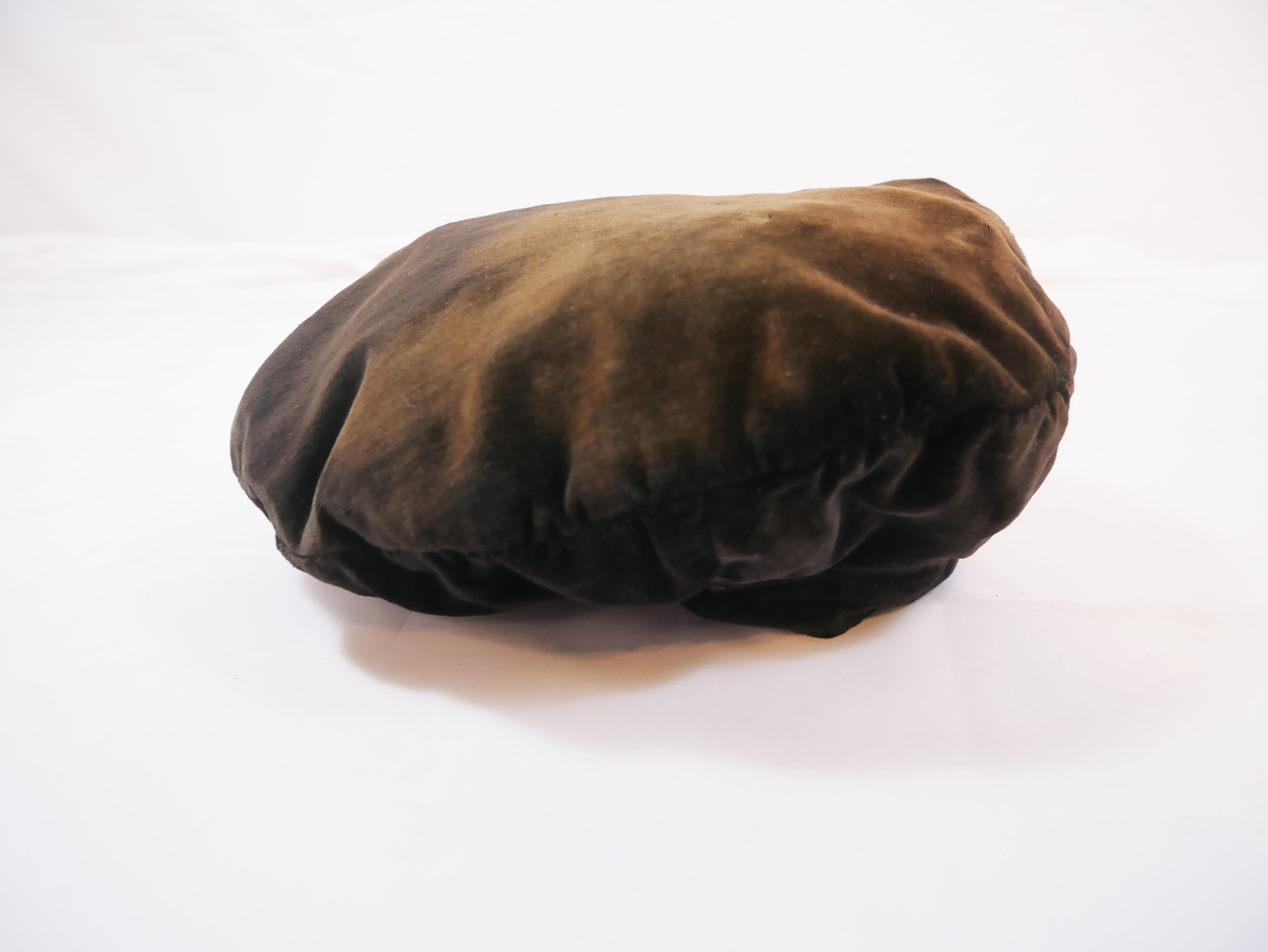 Hovudplagg av  "alpehuetype" med flat, rund pull og linning rundt hovudet. Det er mogleg det her skal vere band eller anna dekor. Fôra med ubleika bomull.