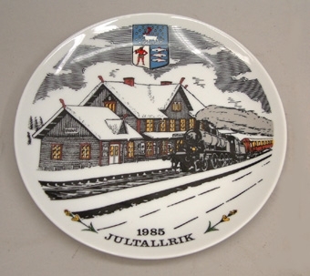 Jultallrik av vitt porslin med dekor i svart, rött och gult.
Motiv av ett ånglok med personvagn framför Vännäs station. Västerbottens landskapsvapen
Text: "1985 Jultallrik".

Tallriken föreställer Vännäs station, byggnaden har 9 st. drakhuvuden.
Bodens station är identisk med densamma.
Denna utgåva är nr.1.