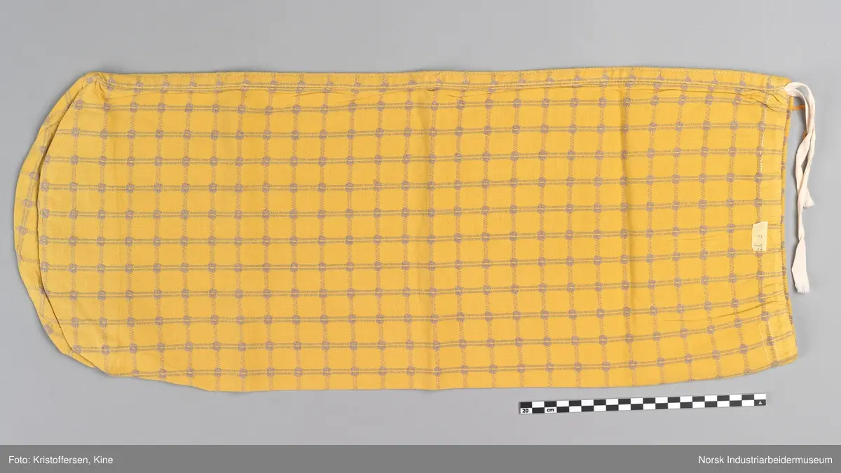 Tekstilpose. Det er søm i den ene siden av posen. Sømmen er en indresøm, det vil si ingen synlige falder og ikke noe stoff som kan trevles opp eller løse tråder på innsiden av posen. Bunnen er sydd av et rundt stoffstykke. Den er festet til posen med en vanlig søm, og kantene er kastet over for hånd. Det er løpegang oppe med bendelbånd gjennom. Dette kan ha vært et trekk til en pute, for eksempel en nakkepute, en pute som ligger i kortendene på en sofa eller seng som res opp om dagen. Det sitter en merkelapp på innsiden av sømmen i øverste kant. Det er en hvit lapp med rød skrift (maskinbroderi). Elektriker. Ovenfor dette merket er det sydd noen få, ujevne sting med beige brodergarn. Dette kunne ha vært en reparasjon, men det er ingen synlige skader i stoffet som skulle tilsi noe slikt. Det kan også være en vaskerimerking, eventuelt rester av en vaskerimerking. Det er også feste en liten, oransje tråd i kanten oppe. Det finnes noe liknende på andre av tekstilene – noe som kan tyde på at det er en merking som sier noe om avdelingen tekstilet hører til – eller det kan være en vaskerimerking. Det er søm i den ene siden av posen.