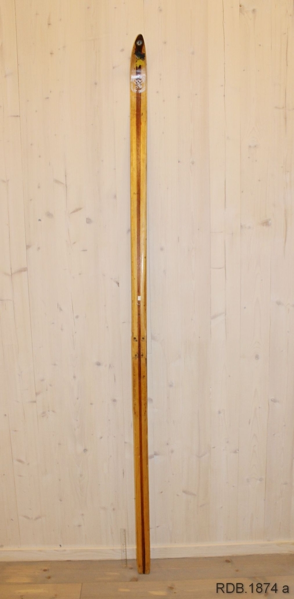 Avsmalende, jevnbred ski med liten bue. Bøy uten tupp. Rett avskåret bakende.
Dekorert med ei brun stripe på trehvit bunn.

Noen stempel fra OL øverst på skiet.