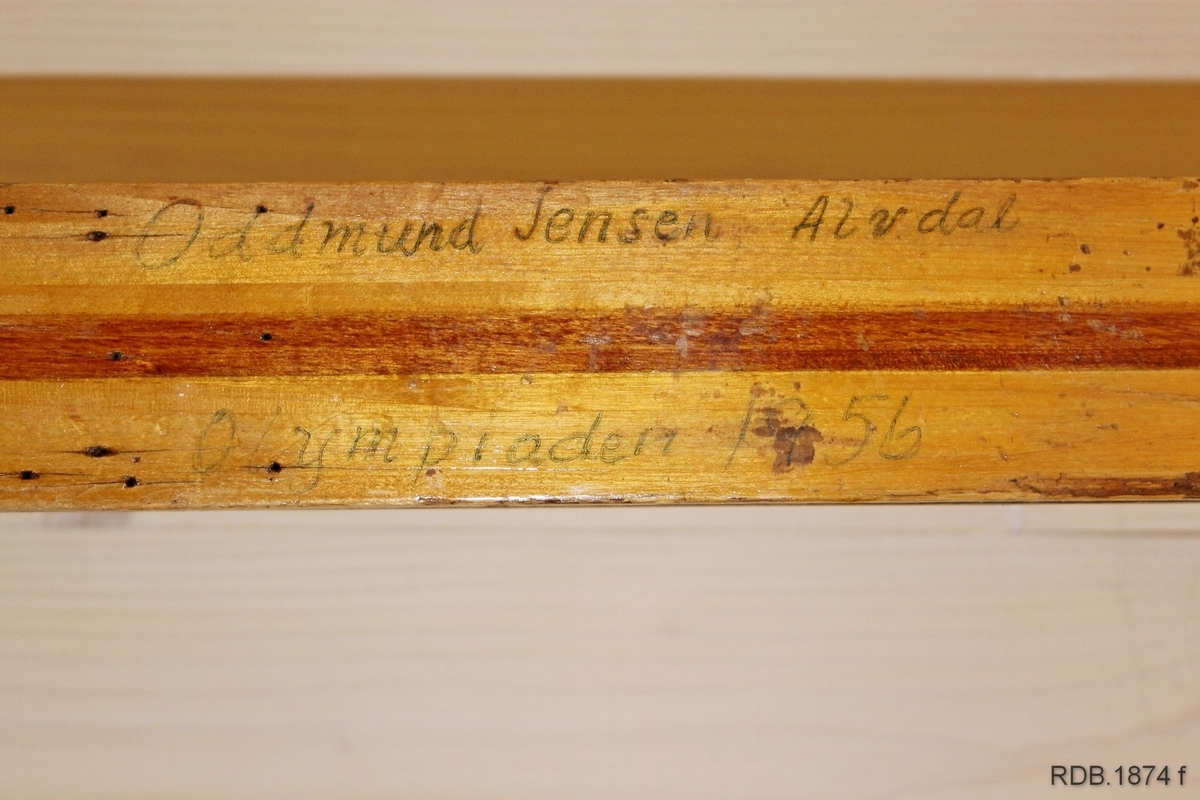 Avsmalende, jevnbred ski med liten bue. Bøy uten tupp. Rett avskåret bakende.
Dekorert med ei brun stripe på trehvit bunn.

Noen stempel fra OL øverst på skiet.