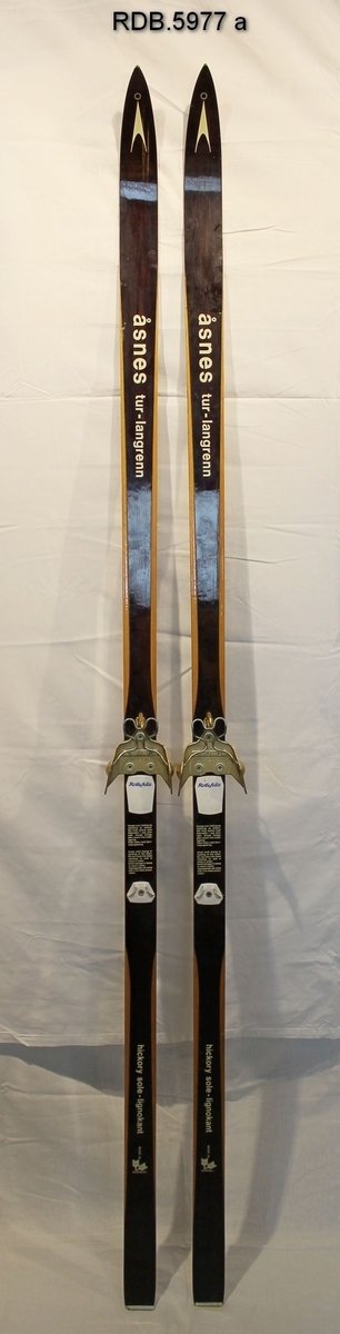 Et par brune, laminerte treski med trekvite kanter, kvit påskrift og dekor. Åsnes-logo på skituppen. Hickorysåle og lignostonekanter. Smal skitupp og stålkanter bak. Påmontert gullfarga rottefellabindinger og to kvite fotplater. Bruksanvisning bak bindingen.