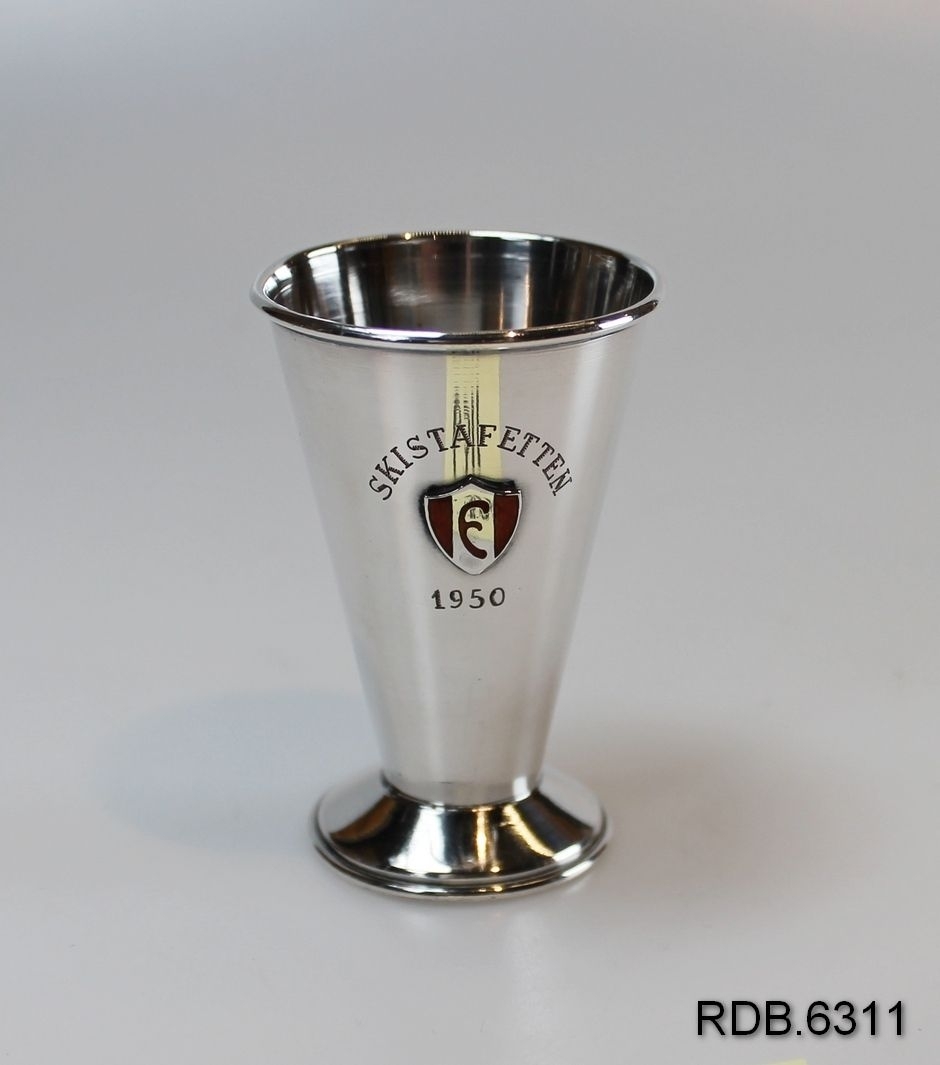 Liten sølvpokal med stett. Foran på pokalen er pålimt et merke/emblem i sølv og rød emalje. Pokalen er noe videre øverst enn nederst.