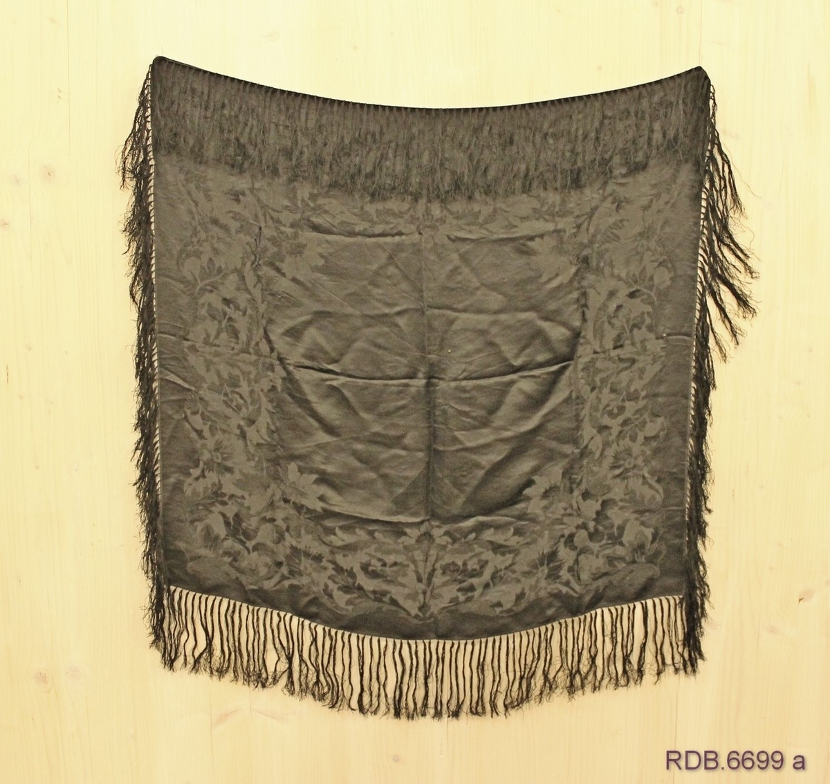 Svart silketørkle vevd i damask med frynser. Bred blomsterbord rundt kanten i stoffet. Jare på to sider og smal fald på de to andre. Frynsene er kytta fast rundt kanten av stoffet.