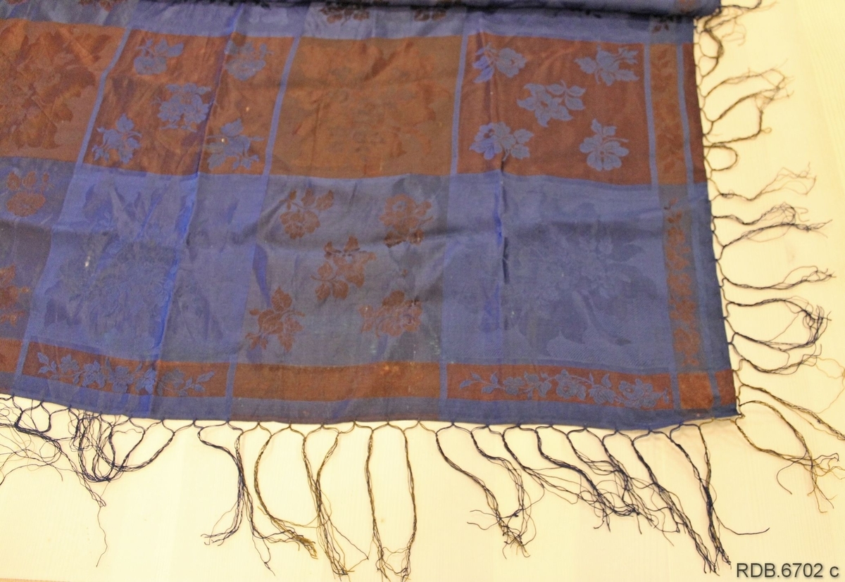 Brunt og blått rutet silketørkle vevd i damask med påknyttede frynser. Jare på to sider og smal fald på de to andre.