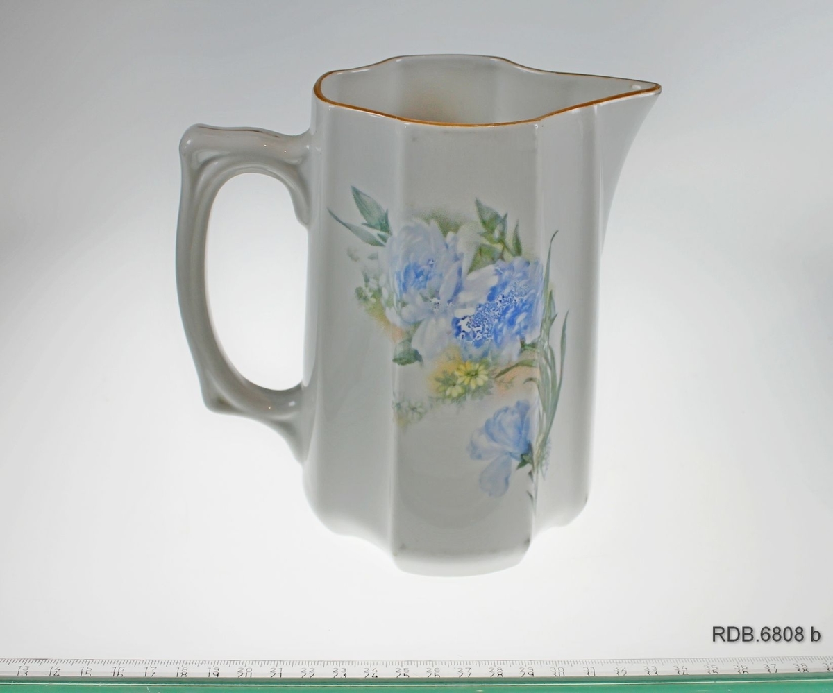 Hvit mugge med stor hank og spiss tut. Åttekantet bunn. Dekorert med trykk av blå og gule blomster og grønne blad på sidene. Gullkant (noe avslitt) på øvre kant og hank. Fabrikkstempel under.