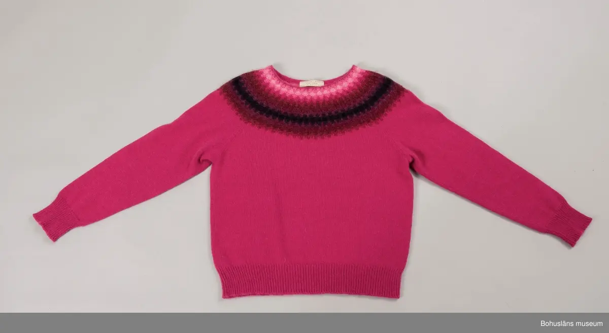 Nyproduktion av stickad jumper med lång ärm, stickad enligt "gamla rundstickningen". Oket är mönsterstickat med räta och aviga maskor i infärgade nyanser av rosa , vinrött, lila och svart. övriga partier slätstickade i rosa. Dubbelvikt halsringning, på baksidan resårstickad. Resårstickade ärmmuddar och nederkant. Fastsydd etikett på insidan halsringningen med vävd text: "BOHUSLÄNS MUSEUM efter original BOHUS STICKNING".