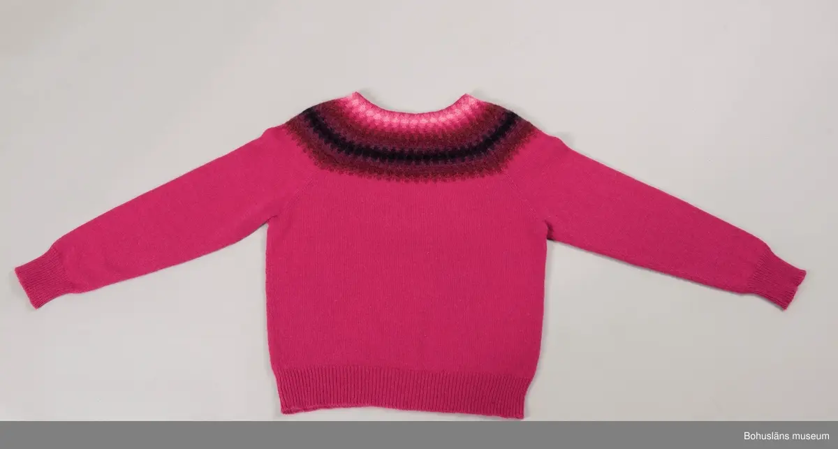 Nyproduktion av stickad jumper med lång ärm, stickad enligt "gamla rundstickningen". Oket är mönsterstickat med räta och aviga maskor i infärgade nyanser av rosa , vinrött, lila och svart. övriga partier slätstickade i rosa. Dubbelvikt halsringning, på baksidan resårstickad. Resårstickade ärmmuddar och nederkant. Fastsydd etikett på insidan halsringningen med vävd text: "BOHUSLÄNS MUSEUM efter original BOHUS STICKNING".