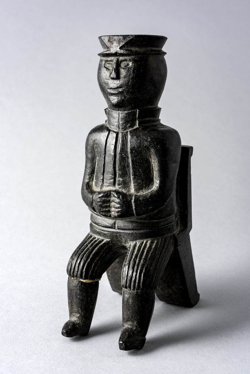 Liten steinskulptur som forestiller mann med hatt sittende på stol uten sete. Bokstøtte?
