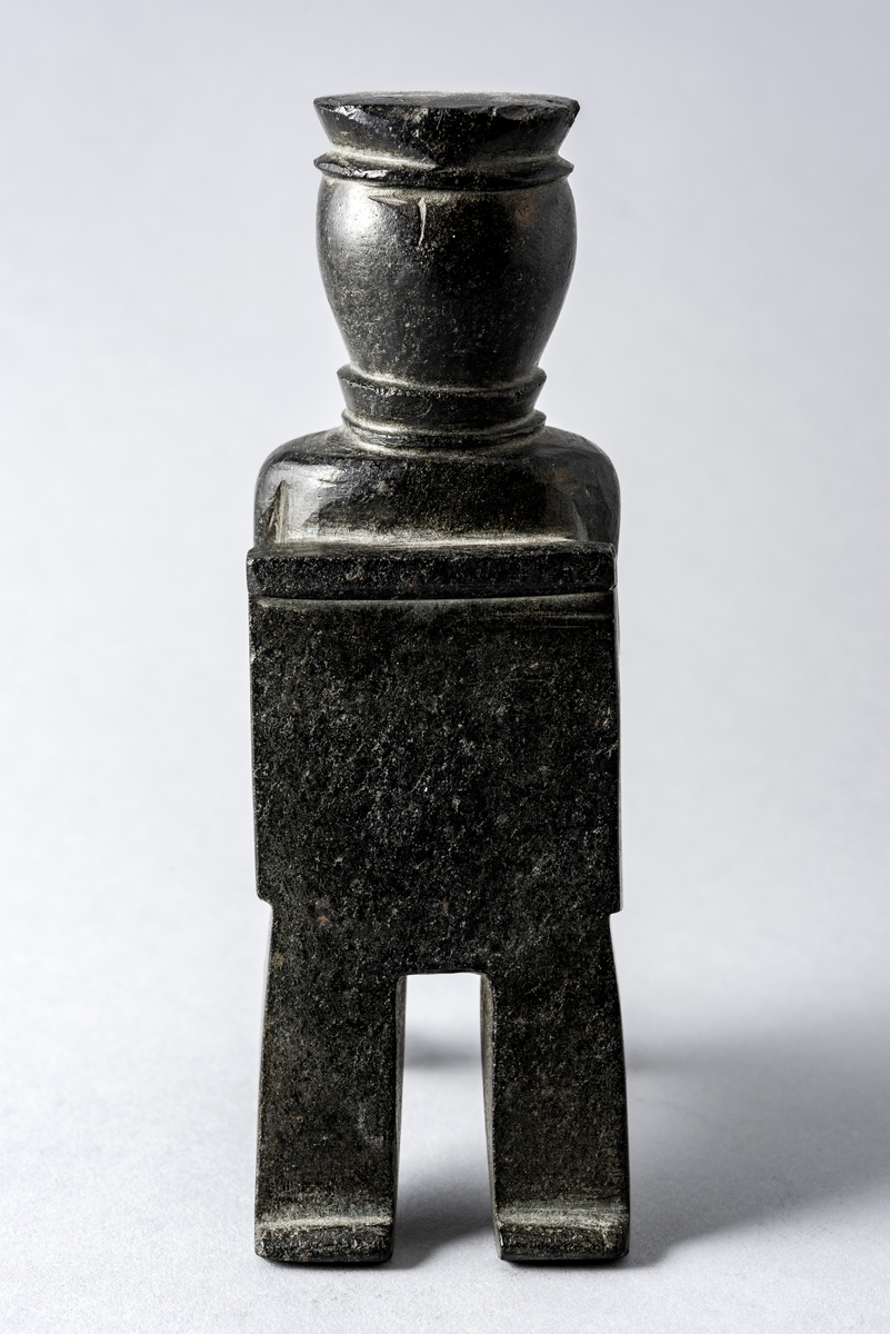 Liten steinskulptur som forestiller mann med hatt sittende på stol uten sete. Bokstøtte?