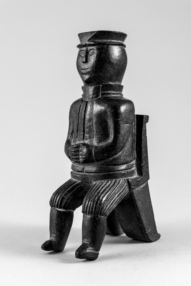 Liten steinskulptur som forestiller mann med hatt sittende på stol uten sete. Bokstøtte?