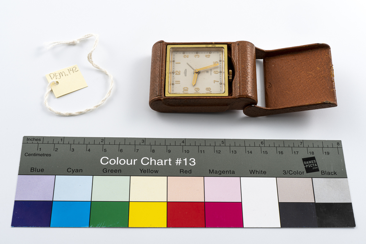 Reiseklokke fra merket Angelus fra Sveits. Ur med i messingfarge festet i brunt skinnetui. Med lokk som i åpen posisjon fungerer som støtte slik at det kan stå på bordet. Uret kan vippes fram for å avdekke baklokk med innstillingsknapper. Har tilhørt Sara Fabricius.