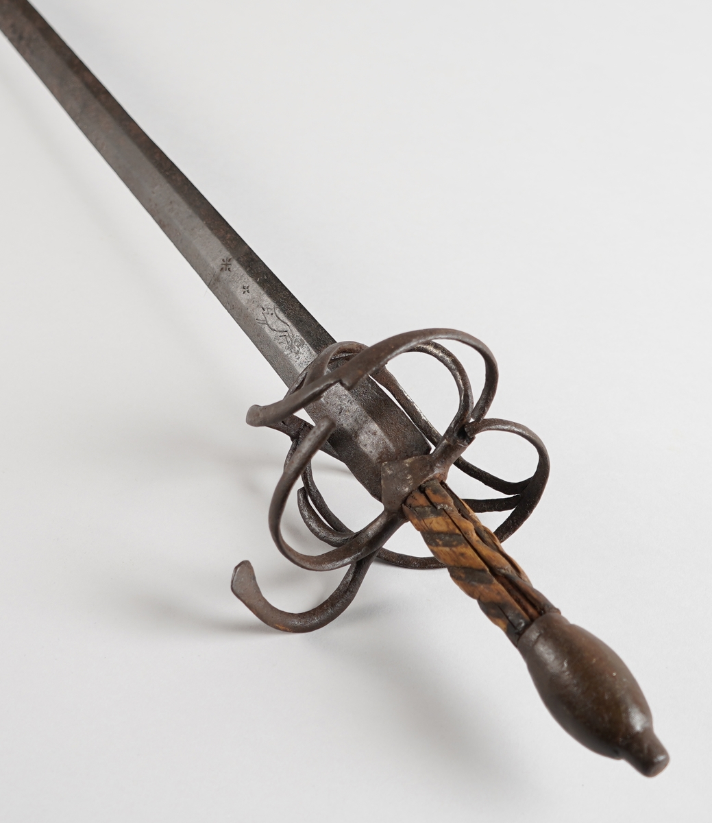 Kårde. Lang, rett klinge med tre kantar (på kvar side). Handtak i tre med spiralliner, solid jarnknapp nedst og metall handbeskyttelse. På kva side av klinga, ned mot handtaket, er det gravert tre kross eller stjerner, og innimellom første og andre stjerne, eit løpande dyr.