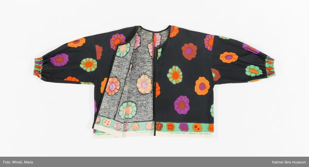 KLM 46157:644. Jacka. För dam. Av svart blommönstrat tryckt tuskaftat bomullstyg. Blommorna är stora, färgglada och strödda över tyget. Rak modell. Utan knäppning, kanter kantade med svart snedslå. Ärm avslutad med insydd resår. Tygets stadkant synlig i jackans nederkant, som en detalj. Tryck i grönt på stadkanten: KATUJI WAKISAKA "NEKKU" © MARIMEKKO OY SUOMI FINLAND 1972.