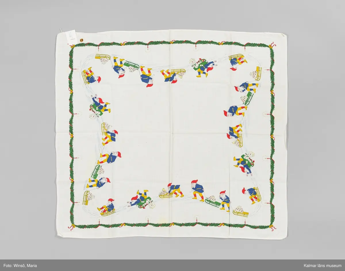 KLM 46157:673. Duk. Julduk. Av vit tuskaftad bomull. Kvadratisk. Tryckt mönster med tomtar som drar eller åker kälke i blått, grönt, rött och gult. Längs ytterkanterna granrisbård med röda band och tända ljus. Fållad i två sidor, de andra två stadkant. 
Fasthäftad vit papperslapp i ena hörnet med handskriven i blå kulspets text: 35-.