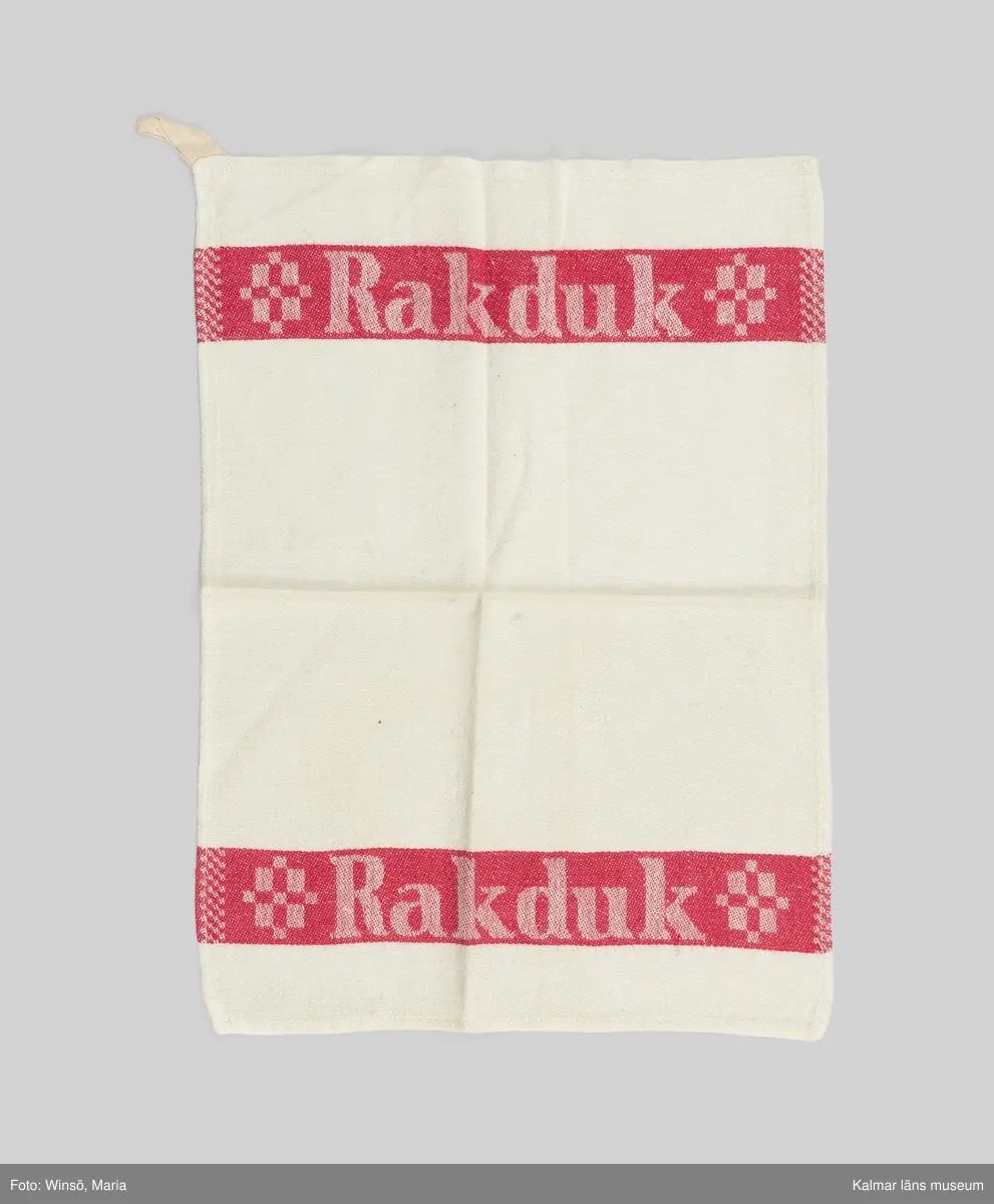 KLM 46157:677. Handduk. Rakduk. Av blekt damastvävt linne. Röda bårder vid kortsidorna med invävd text: Rakduk. Vid mitten motiv vitt i vitt,  rakborste och hyvel. Fastsydd hank i ena hörnet. Maskinfållad längs tre sidor, stadkant vid den fjärde.