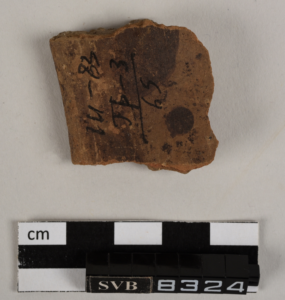 Lett buet keramikkfragment med bruddkanter på tre av sidene. Fragmentet er lys brunt i farge. Fargen i bruddkantene er grå.