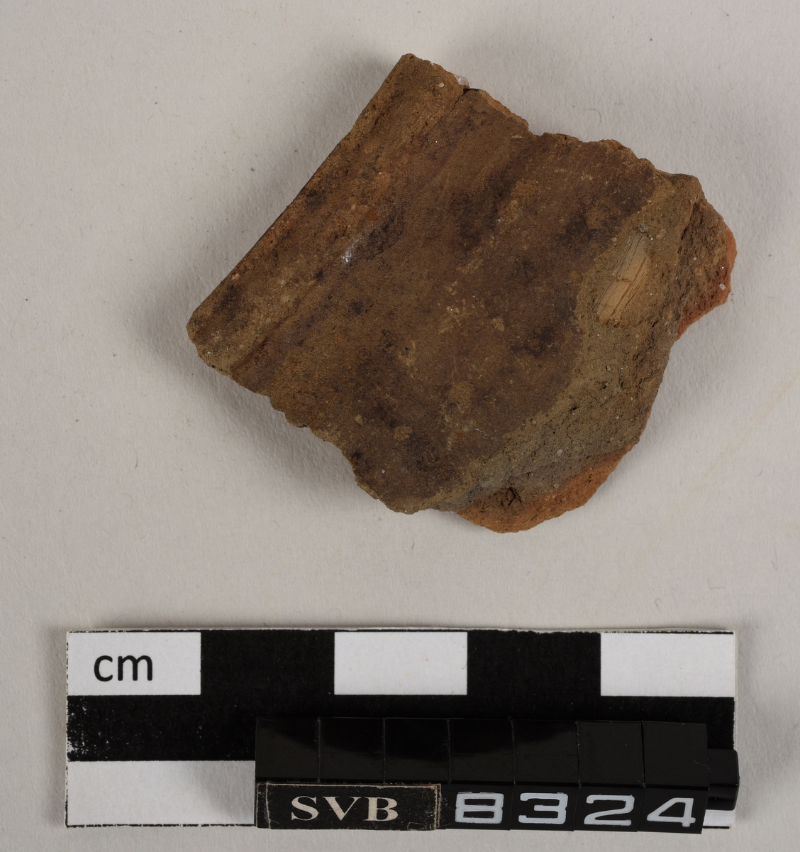 Lett buet keramikkfragment med bruddkanter på tre av sidene. Fragmentet er lys brunt i farge. Fargen i bruddkantene er grå.