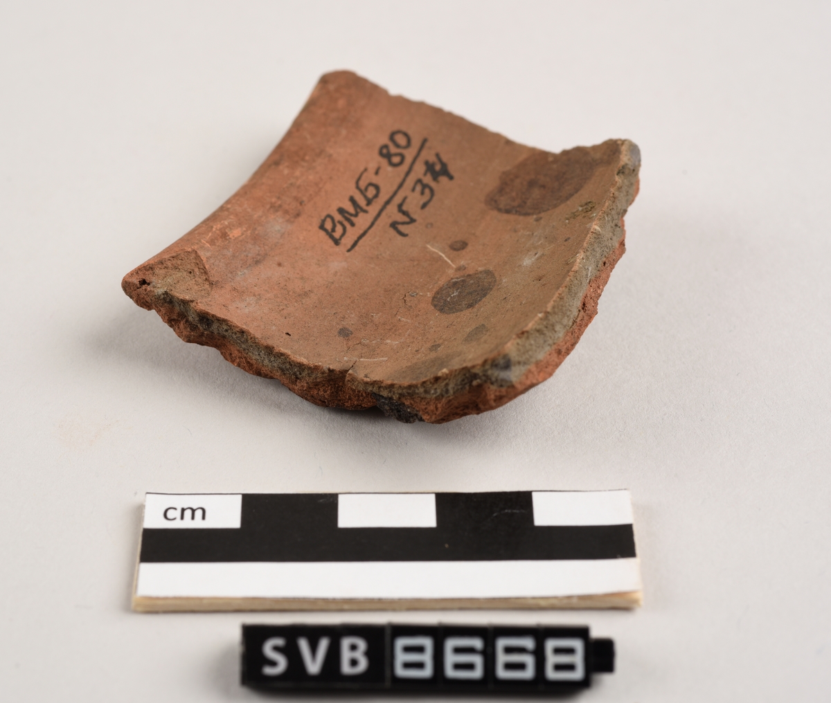 Buet keramikkfragment med bruddkanter på tre av sidene. Fragmentet er rødlig i farge. Bruddkantene er grå i farge.